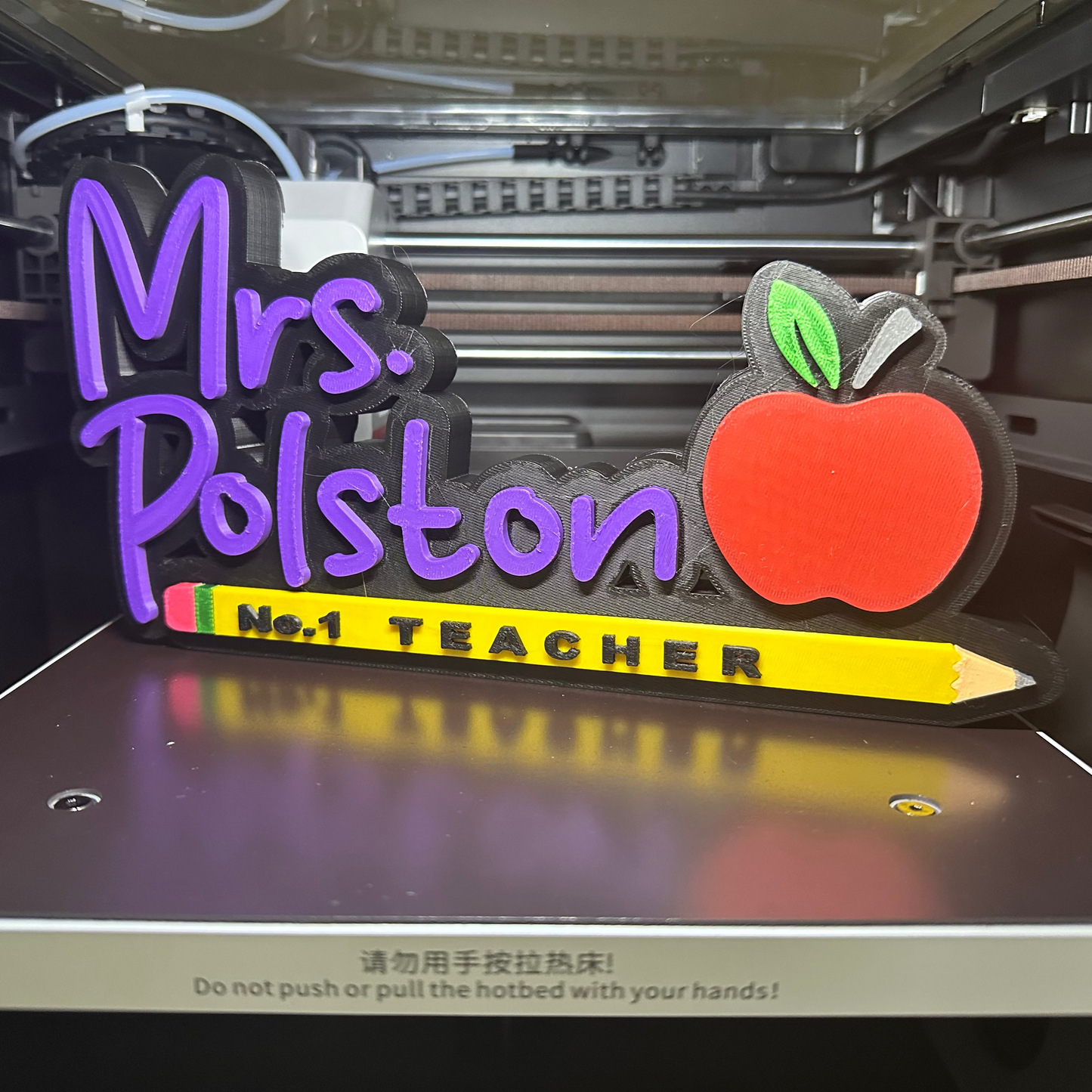 3D-printed nameplate with 'Mrs. Polston No. 1 Teacher' text and apple design on a 3D printer bed.