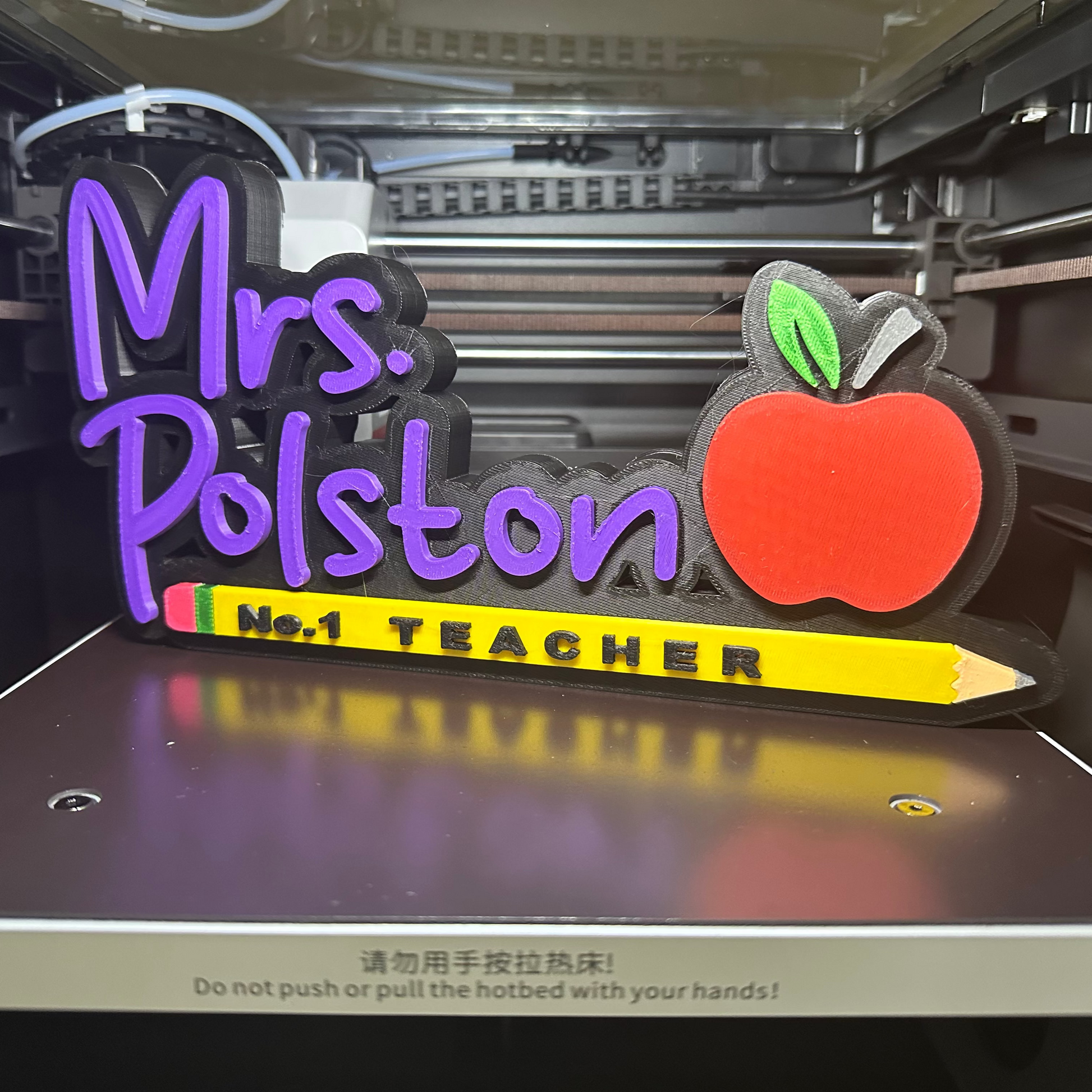 3D-printed nameplate with 'Mrs. Polston No. 1 Teacher' text and apple design on a 3D printer bed.
