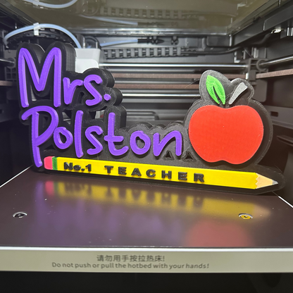 3D-printed nameplate with 'Mrs. Polston No. 1 Teacher' text and apple design on a 3D printer bed.