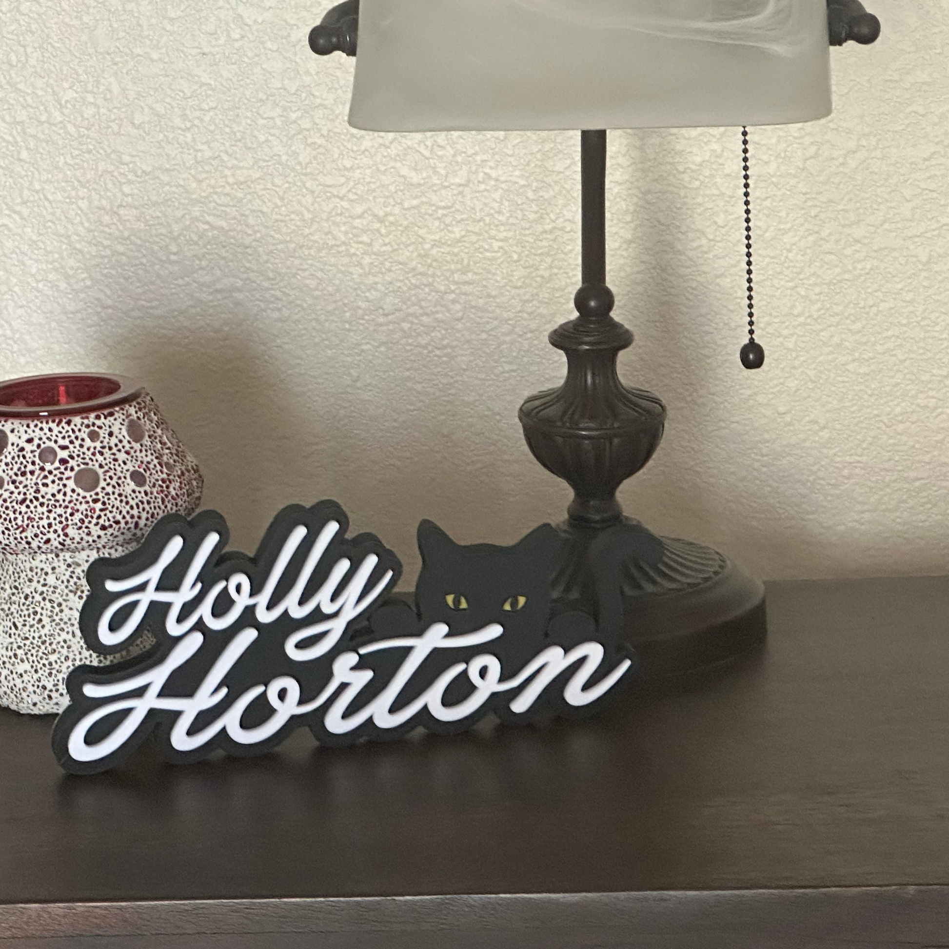 Decorative sign with 'Holly Horton' on a surface with a lamp and small lantern in the background.
