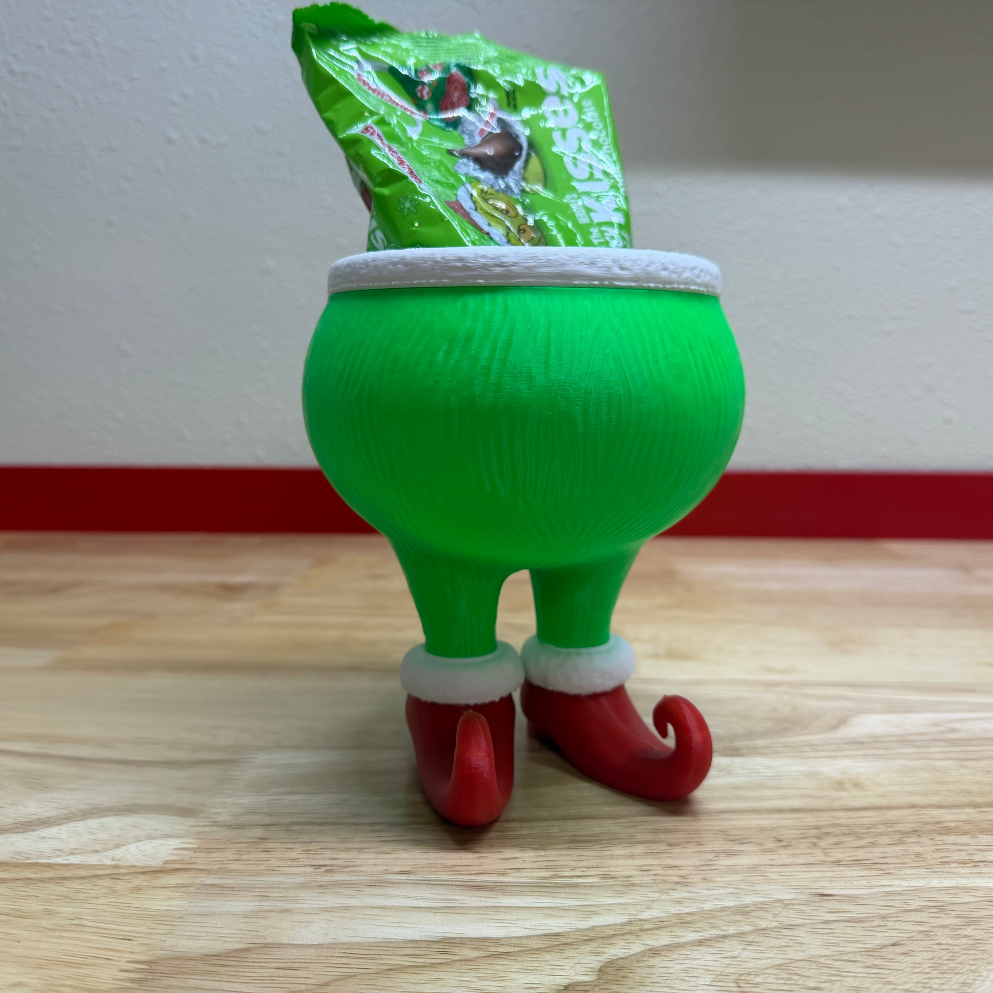 Green elf-shaped container with a white rim and red feet on a wooden surface.