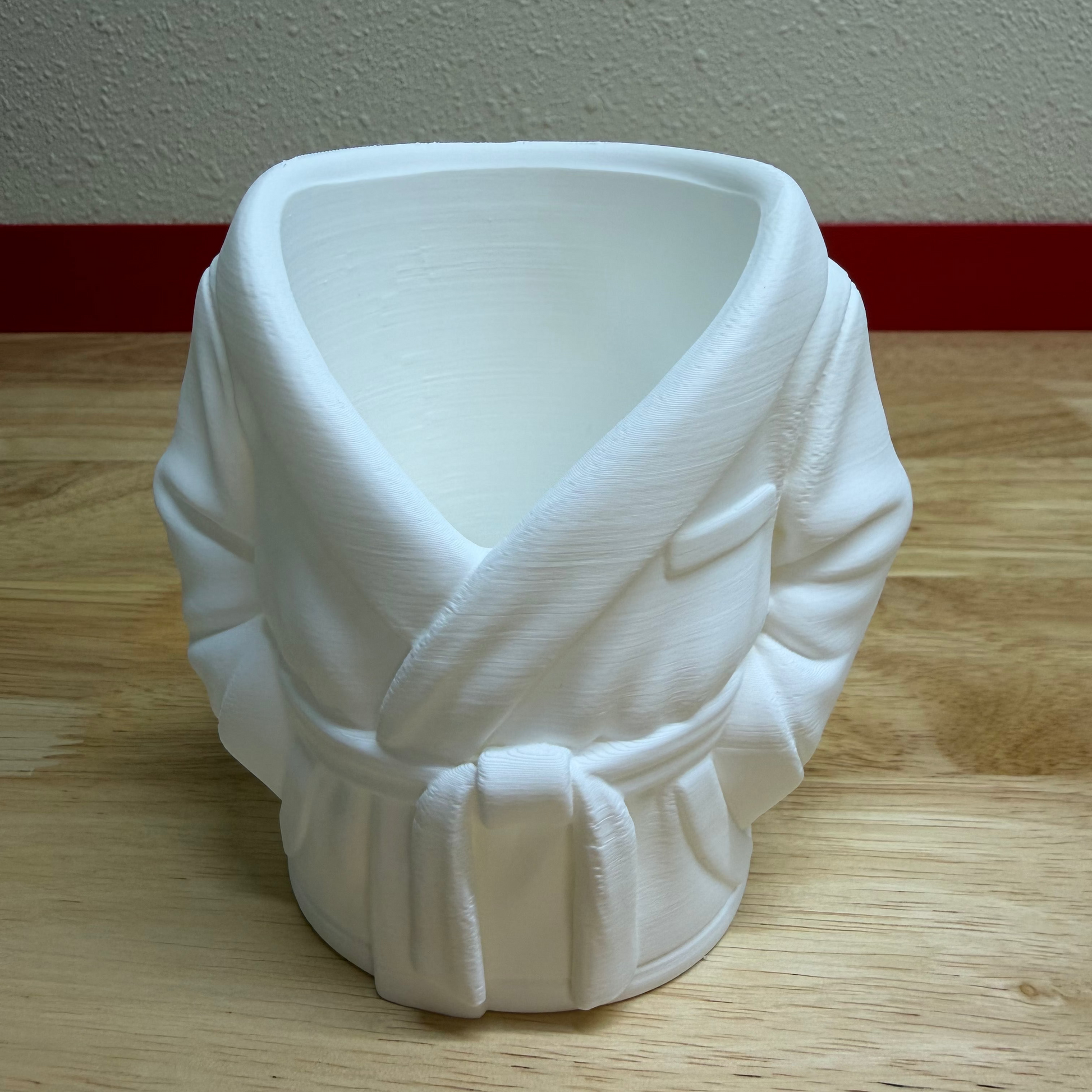 White ceramic pot shaped like a robe on a wooden surface with a plain wall background