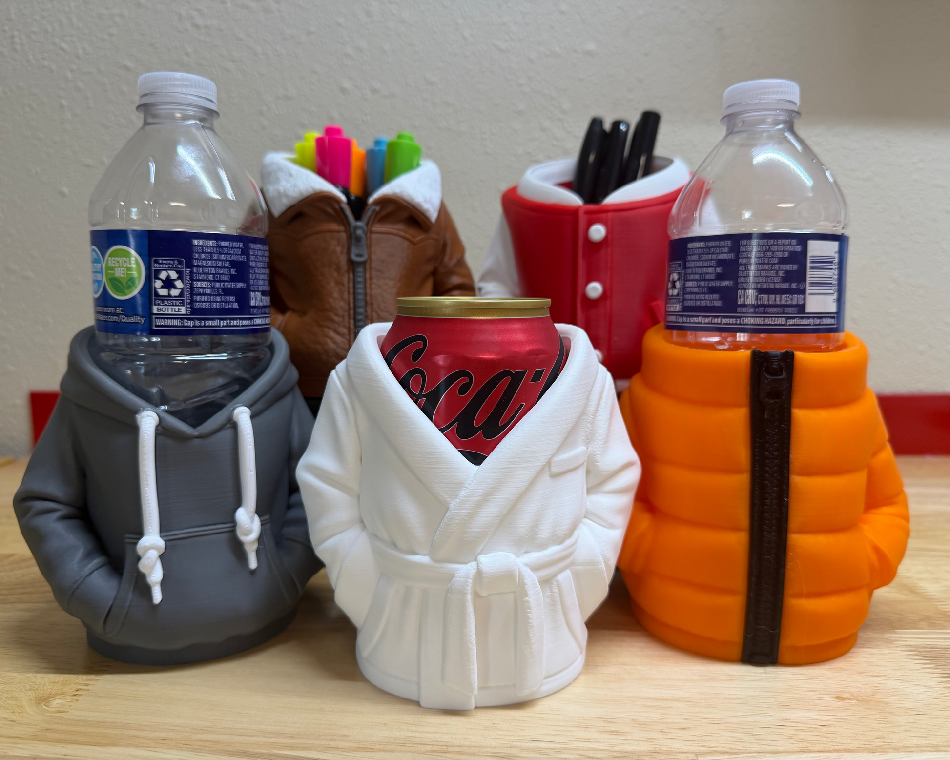 Three novelty drink coolers shaped like a hoodie, robe, and grenade on a wooden surface.