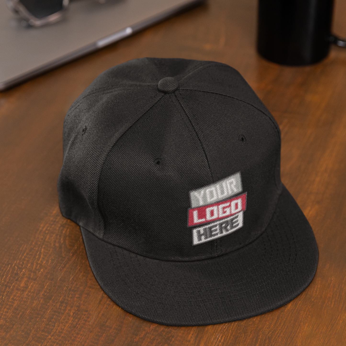 Black cap with 'Your Logo Here' text on a wooden surface with sunglasses and a mug.