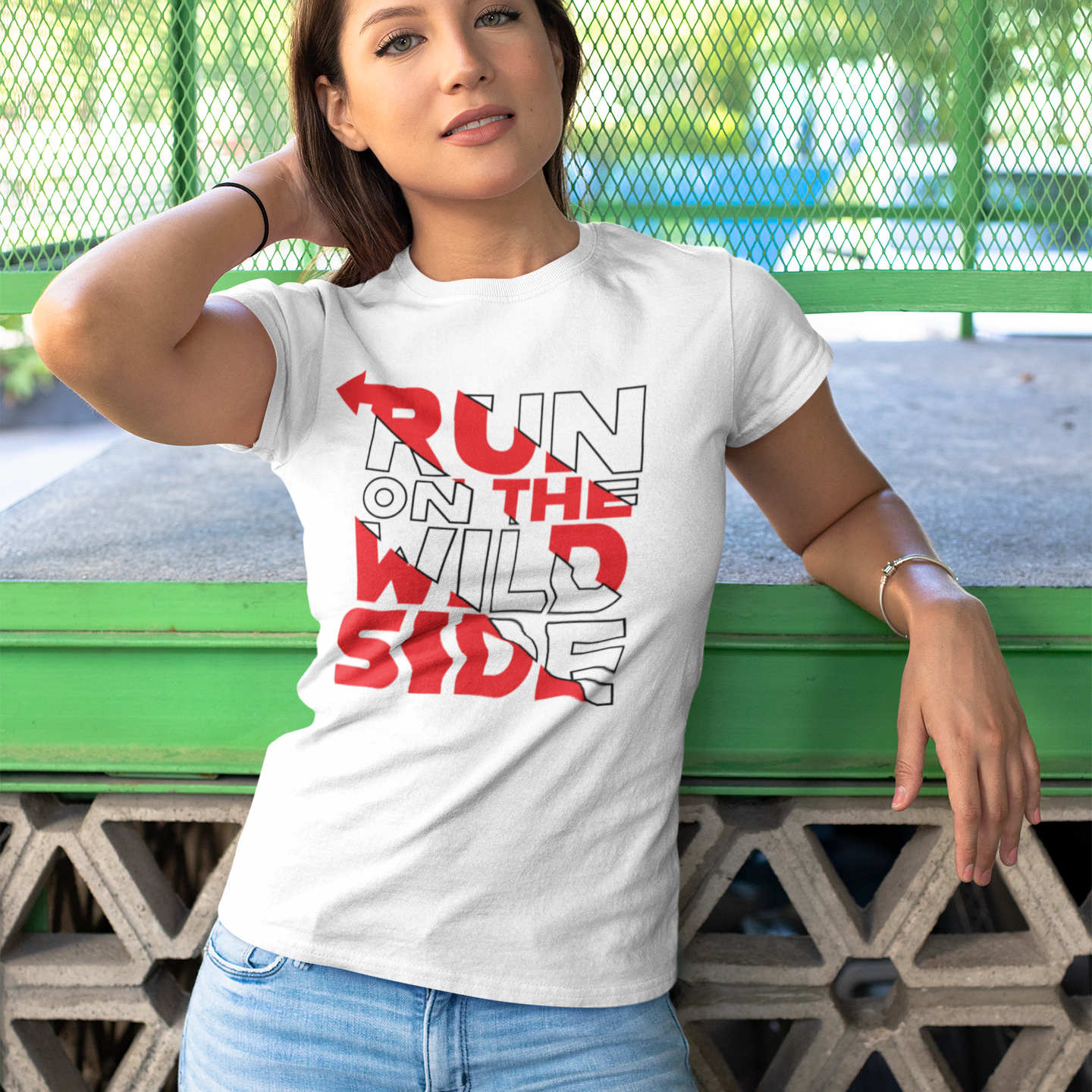 Woman wearing a white t-shirt with 'Run on the Wild Side' text, standing against a green metal fence.